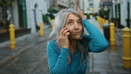 Fototapeta premium Senior woman with grey hair chatting on a phone in a vibrant urban street with yellow bollards and a backdrop of city architecture.