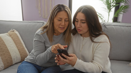 Mother and daughter sitting together on a comfortable sofa in the living room, looking at a smartphone, capturing a moment of bonding and technology use in a warm home environment.