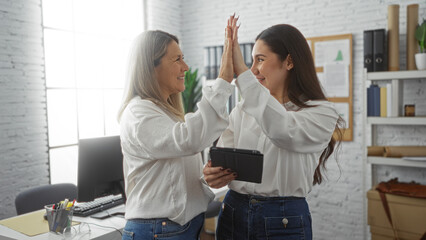 Women colleagues high-five cheerfully in a modern office, using a tablet, symbolizing teamwork and success in a collaborative workplace environment.