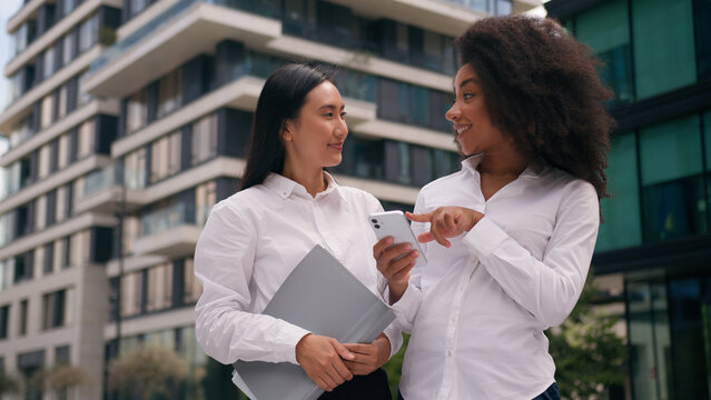 African American Asian Korean women females colleagues businesswomen business using mobile phone smartphone online discuss talking social media internet ebusiness employees city outdoor communication