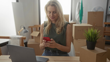 Woman smiling indoors with laptop and smartphone in living room of new home surrounded by boxes...