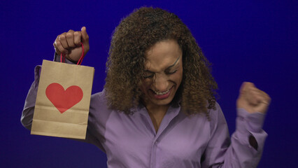 Woman holds shopping bag with heart graphic, smiles broadly and raises fist in studio; excitement...