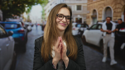 Young woman with brown hair wearing glasses clasping hands with a gentle smile in a busy city...