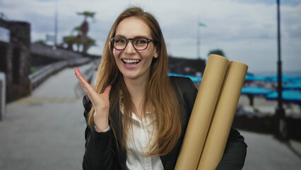 Smiling woman wearing glasses holds two cardboard tubes and touches cheek on street by the beach...
