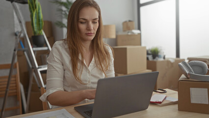 Woman typing on laptop amid unpacked cardboard boxes at wooden table in new home building; focus...