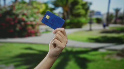 Man holding blue credit card in hand against blurred park background showcasing green grass and colorful flowers under sunlight with a clear sky.