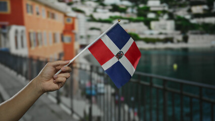 Hand holding dominican republic flag by seaside with blurred city background.