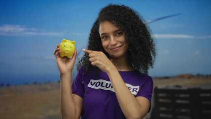 Woman smiling in field holds piggybank against windmill backdrop, showcasing volunteer spirit and...