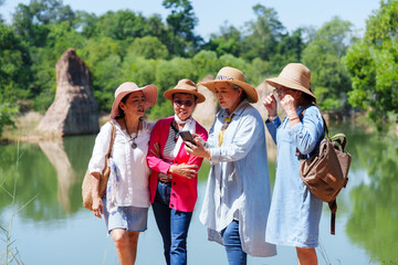 Group senior Asian women standing together looking smartphone laughing casual colorful clothing hats backpacks lakeside outdoor enjoying natural environment bright sunny day happy retirement trip