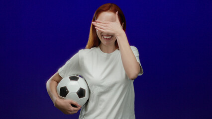 Woman holding soccer ball smiles playfully against a vibrant blue background, showcasing her red hair and casual white shirt in a vivid and energetic sports theme setting.