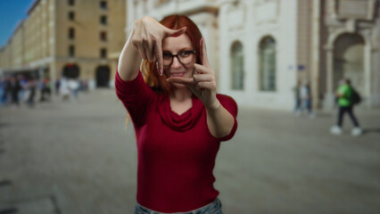 Fototapeta premium Woman with red hair and glasses forms frame with fingers while smiling on city street, expressing creativity and joy in bustling urban outdoor setting.