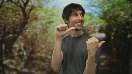 Man in gray shirt thumbs pointing to chest on a sunlit green forest path during daytime; self...
