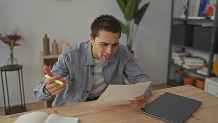 Young man analyzing document at home, sitting in modern living room with laptop and books, conveying focus and concentration, surrounded by stylish decor and personal items.