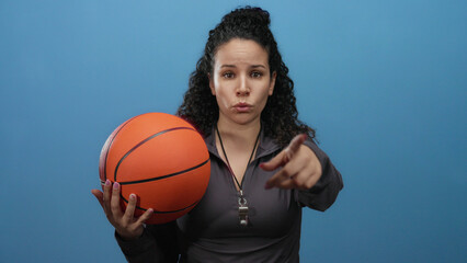 Woman coach holding basketball and pointing against blue wall with determined expression on...