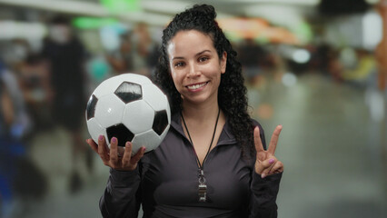 Woman holding soccer ball and making peace sign in an airport terminal, symbolizing sports and...