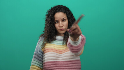 Woman gesturing no with hand towards camera against isolated green background, wearing colorful...