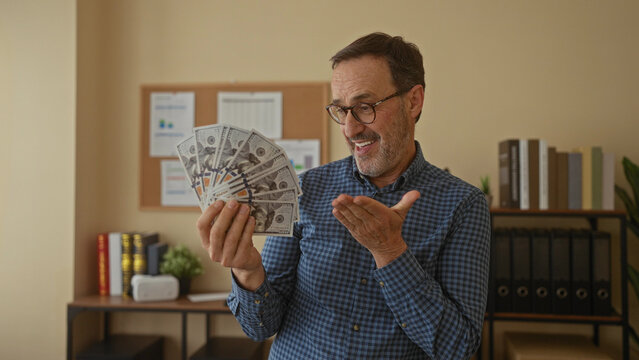 Man holding cash indoors in an office setting with a joyful expression, surrounded by books and plants, showcasing financial success and happiness at work.