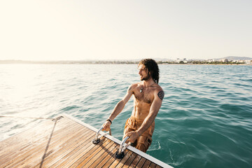 Fototapeta premium Shirtless young man with long hair getting out of the sea, climbing aboard a sailboat using a ladder at sunset