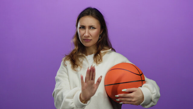 Young woman holding basketball against purple background with a perplexed expression, isolated and wearing casual white hoodie.