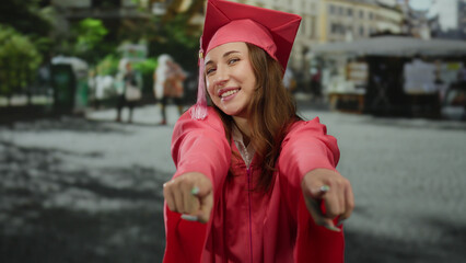 Young woman in pink graduation gown celebrates with pointed fingers on a bustling urban street...