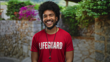African american man lifeguard smiling outdoors wearing red shirt against vibrant flowered...