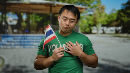 Young man wearing green shirt holds thai flag in park setting, emphasizing volunteer spirit and...
