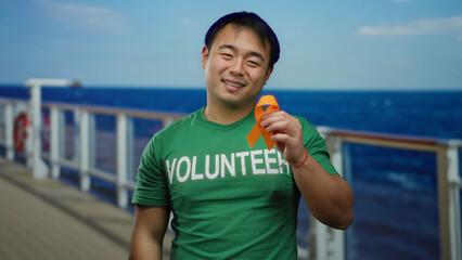 Young man wearing volunteer shirt holds orange ribbon on cruise ship with ocean background...