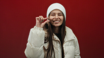 Woman in white coat and beanie gesturing on red background exudes warmth and style while expressing...