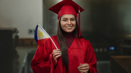 Young woman in a red graduation gown holding a philippines flag smiles proudly indoors, symbolizing...