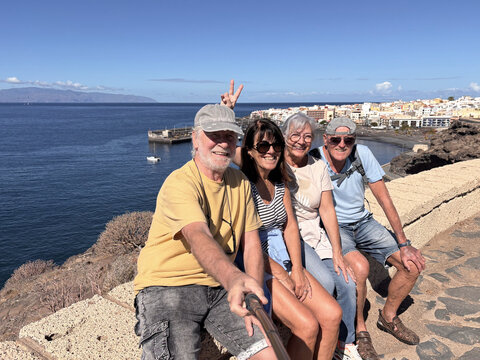 Cheerful group of four seniors on outdoor hike at the edge of the sea sitting taking a picture with a selfie stick. Relaxed and serene retirement. Horizon over the water and view of La Gomera island
