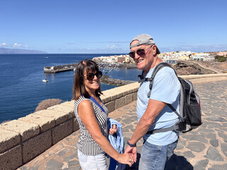 Cheerful senior couple hand in hand on outdoor hike along the sea enjoying sunny day in good...
