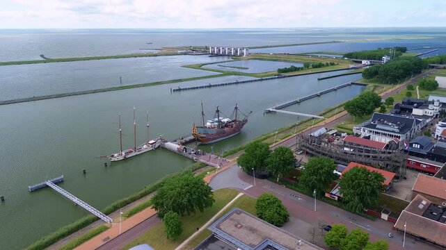 An aerial view of a coastal town with a harbor, colorful buildings and a replica ship docked along the water. Surrounding areas include parking lots and green spaces. - Powered by Adobe