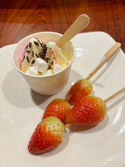 Vanilla soft serve with chocolate sprinkles and marshmallow, and strawberry on skewer, served on a white plate.