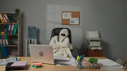Halloween businessman ghost in white sheet sits at office desk works on laptop, sad expression...