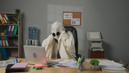 Halloween businessman ghost in white sheet sits at office desk works on laptop, sad expression...