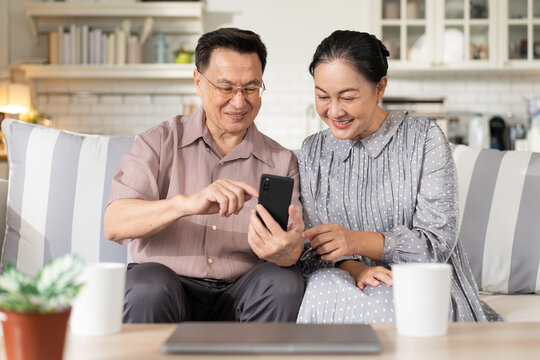 Asian senior couple enjoy using mobile phone on sofa. grandparent learning and connecting with social media, video call with family, shopping online, surfing the internet. - Powered by Adobe