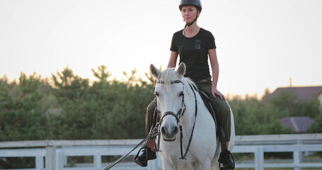 Female equestrian riding a majestic white horse in a tranquil manege, enjoying a serene sunset...