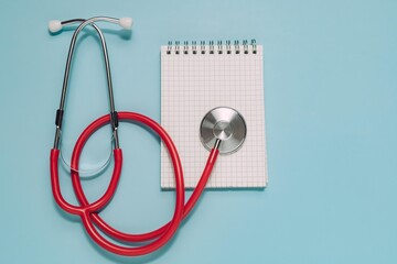 Vibrant red stethoscope next to an open spiral notepad with grid paper, all on light blue. Ideal for medical consultation, patient records, diagnosis, and healthcare planning.