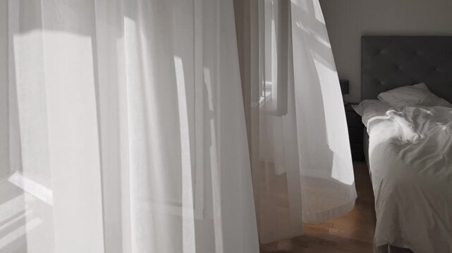 White curtain flying by the wind next to an unmade empty bed. Cozy morning vibes, fresh air coming through the open window. Soft morning sunlight casting shadows on wrinkled cotton bedsheets.