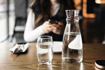 A carafe of water and a glass against the background of a woman in a restaurant.