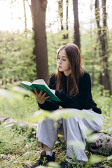 Teenage girl reading a book while sitting on a log in the forest, copy space.