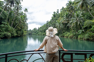 A woman wearing a straw hat glides gracefully on a boat down a serene tropical river. Lush greenery...