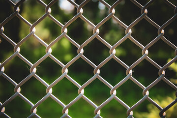 Fototapeta premium close-up view of a chain-link fence set against a blurred green landscape background. abstract texture and concept of barrier or security. field surround with dappled sunlight.