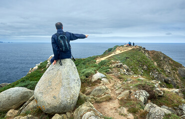 A hiker sitting on a large oval stone points to people near the last rocks at Estaca de Bares cape.