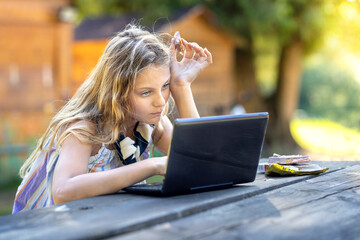 Bored young girl learning with Dubai chocolate in mouth and laptop on wooden table at sunset. Business laughing woman communicating at homework via wifi on computer. Watching film