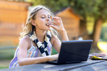 Joyful young girl learning with Dubai chocolate in mouth and laptop on wooden table at sunset. Business laughing woman communicating at homework via wifi on computer