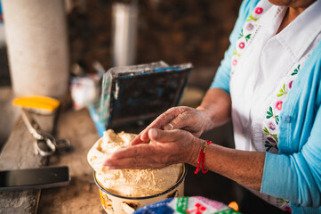An indigenous Mexican woman wearing a white embroidered blouse and blue sweater is preparing the dough with her hands to make tortillas, a traditional Mexican dish.
