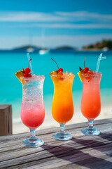 Three vibrant cocktails sit on a wooden table, showcasing pink, orange, and red hues. The tranquil beach and turquoise water create a relaxing atmosphere under the bright sun