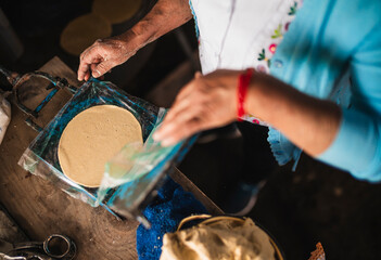 An indigenous Mexican woman wearing a white embroidered blouse and blue sweater, pressing dough into a tortilla press to make tortillas, a traditional Mexican dish.
