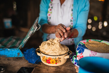 An indigenous Mexican woman wearing a white embroidered blouse and blue sweater is preparing the dough with her hands to make tortillas, a traditional Mexican dish.
